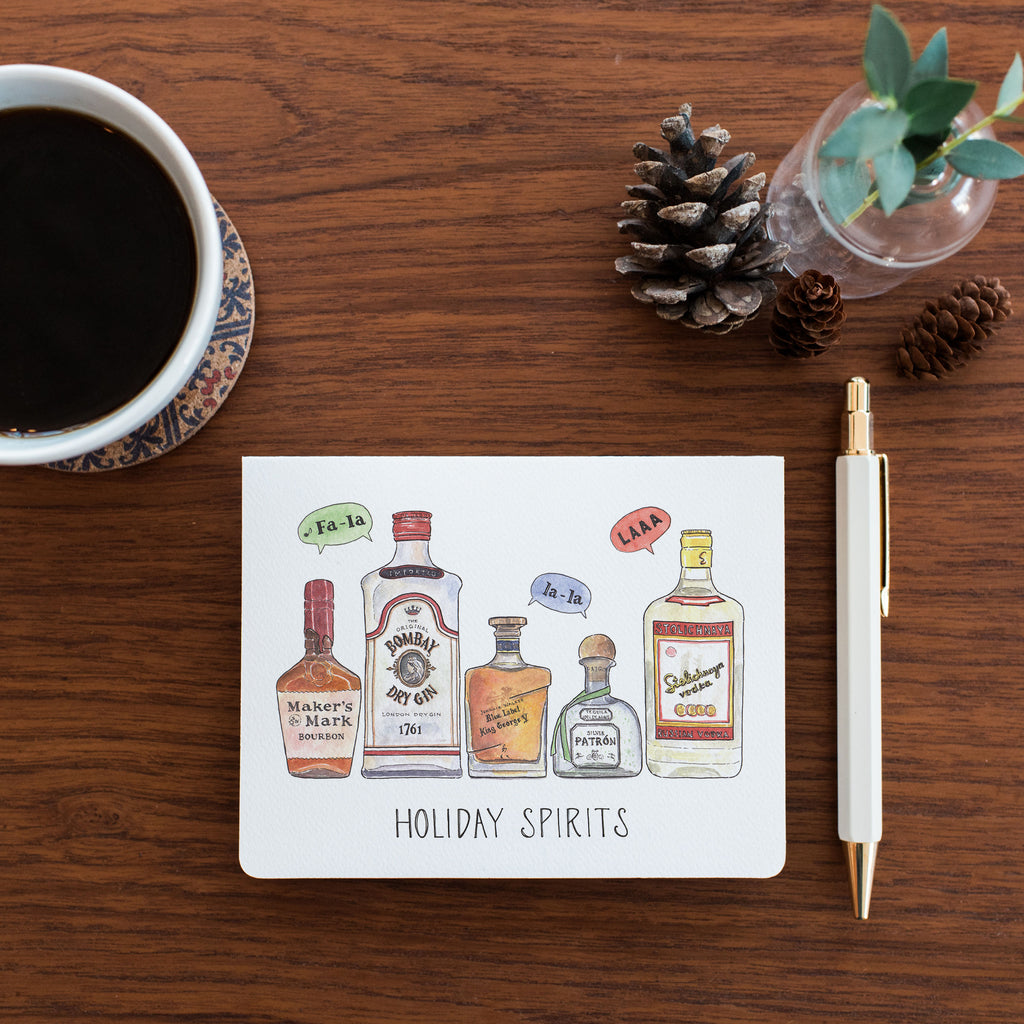Greeting Card with 'Holiday Spirits' illustration on a wooden table with coffee, pinecones, and a pen.