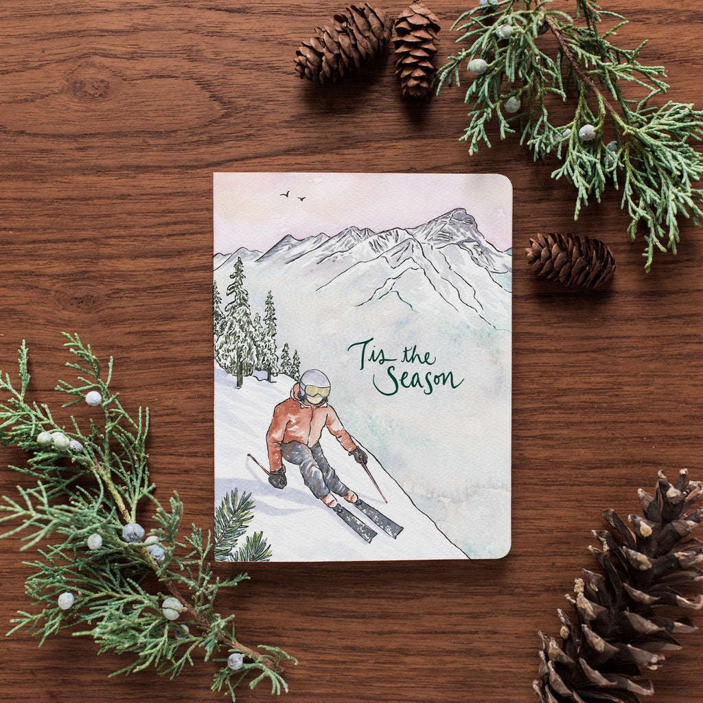Card with skier illustration and 'Tis the Season' text on a wooden surface with pine cones and branches.
