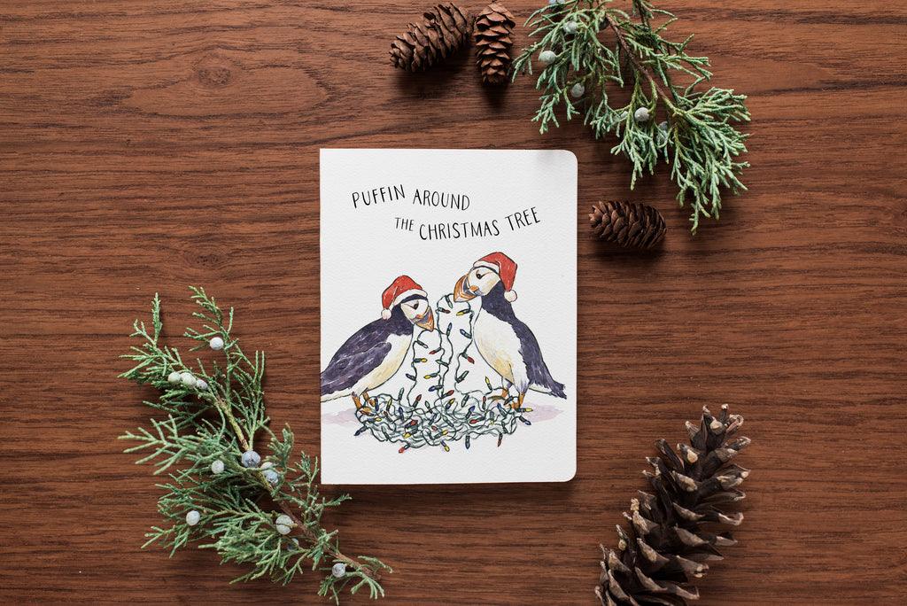 Card with puffin illustration on a wooden surface with pine branches and cones.