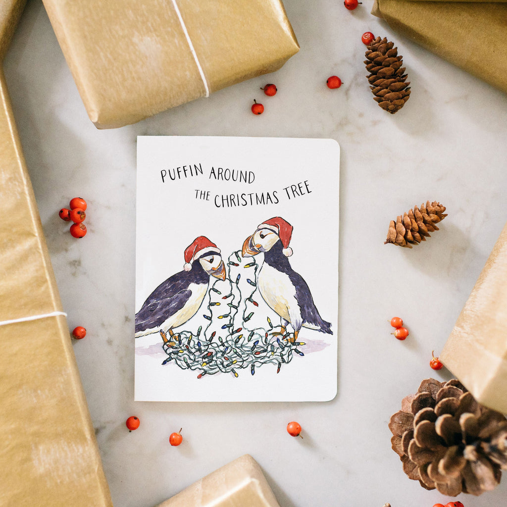 Gifts wrapped in brown paper with a Christmas card featuring penguins and holly leaves on a light gray surface.