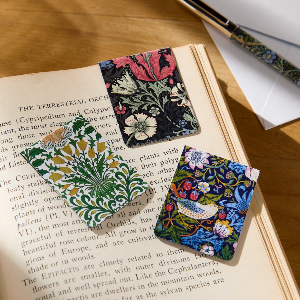 A set of three magnetic bookmarks, featuring some of WIlliam Morris' well-loved antique floral patterns. They are displayed on an open book for scale.