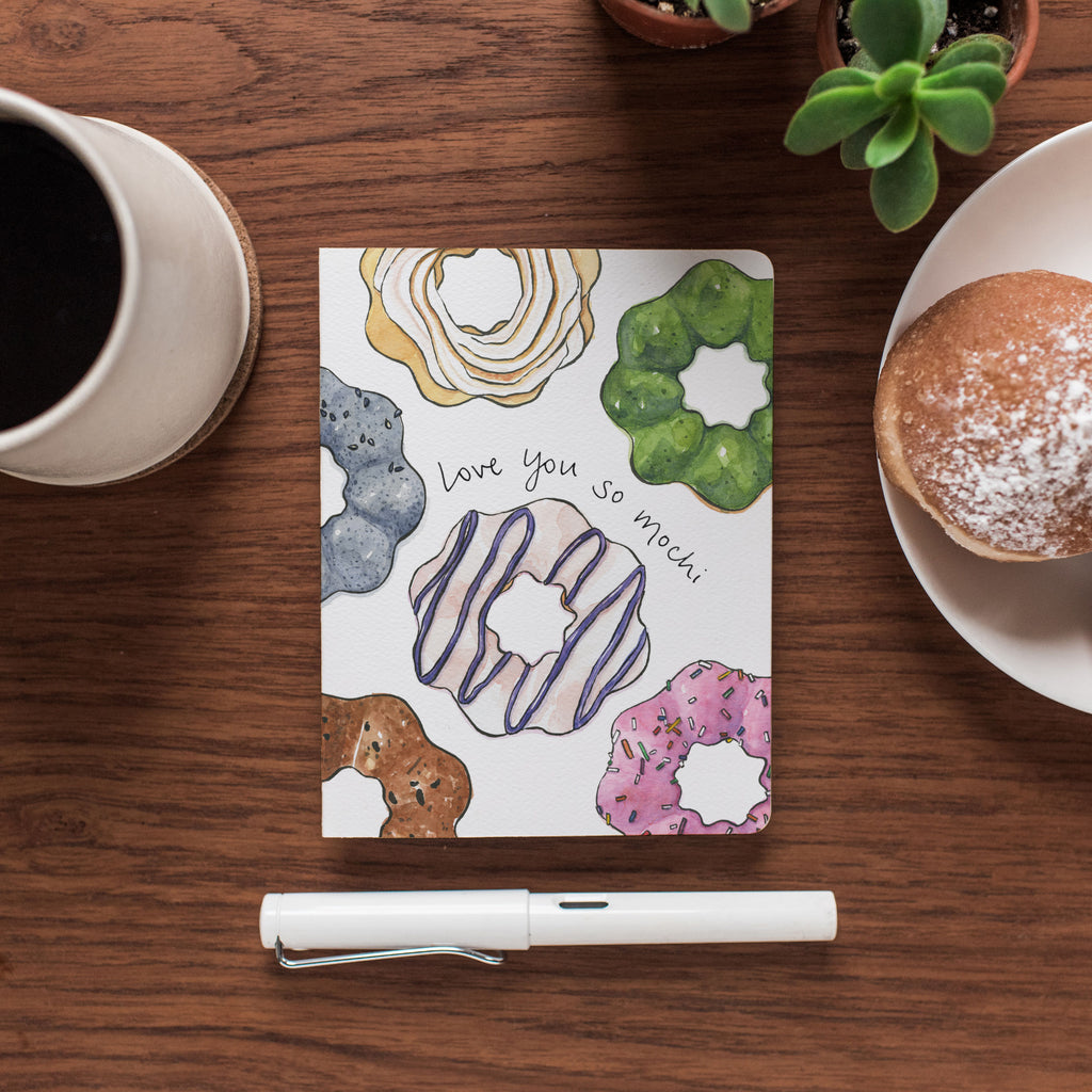 The Mochi Love card rests on a wooden desktop surrounded by a cup of coffee and a donut.