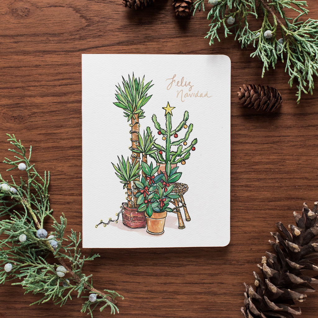 The Feliz Navidad card rests on a wooden table surrounded by pinecones and holly.