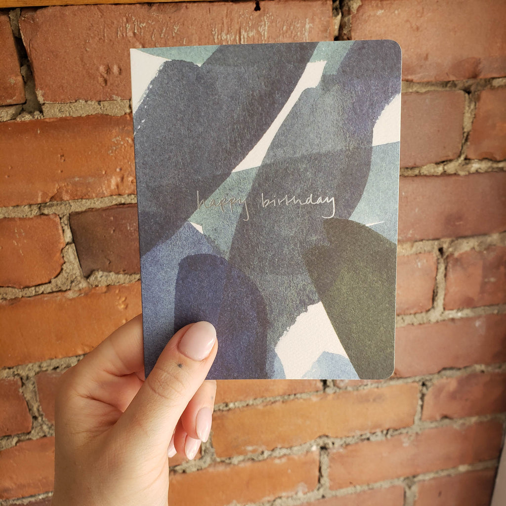 A hand hold the Blue in Green birthday card in front of a brick wall.