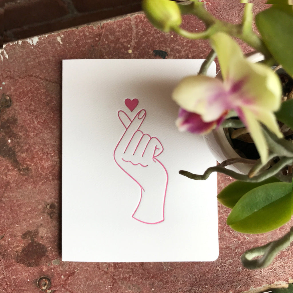 Card front features a letter-pressed fuchsia hand design. The hand makes the popular fingerheart symbol, crossing the thumb and pointer finger to make a heart.